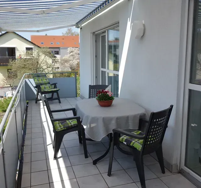 Balkon mit Tisch, Stühlen und Markise. Weißes Gebäude, Blick auf Häuser und Bäume.
