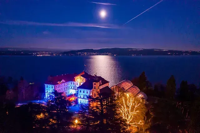 Das Schloss erstrahlt nachts in blauen und rosa Lichtern, über dem See steht der Vollmond.