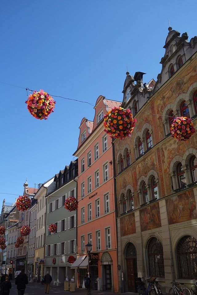 Bunte Gebäude säumen eine sonnige Straße, darüber hängen Blumenkugeln.
