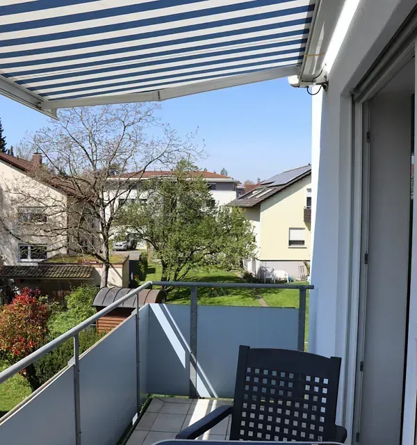 Balkon mit gestreifter Markise, Stuhl, Glasgeländer und Blick auf Häuser und Bäume an einem sonnigen Tag.