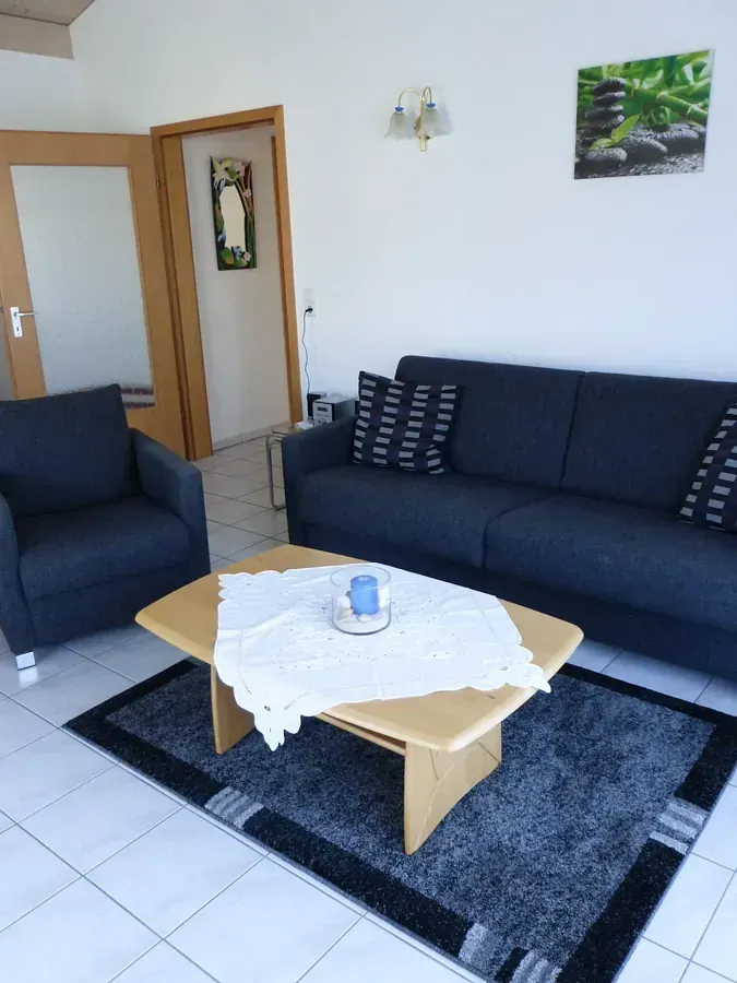 Wohnzimmer mit blauem Sofa, Sessel, Teppich und hölzernem Couchtisch. Türöffnung sichtbar.