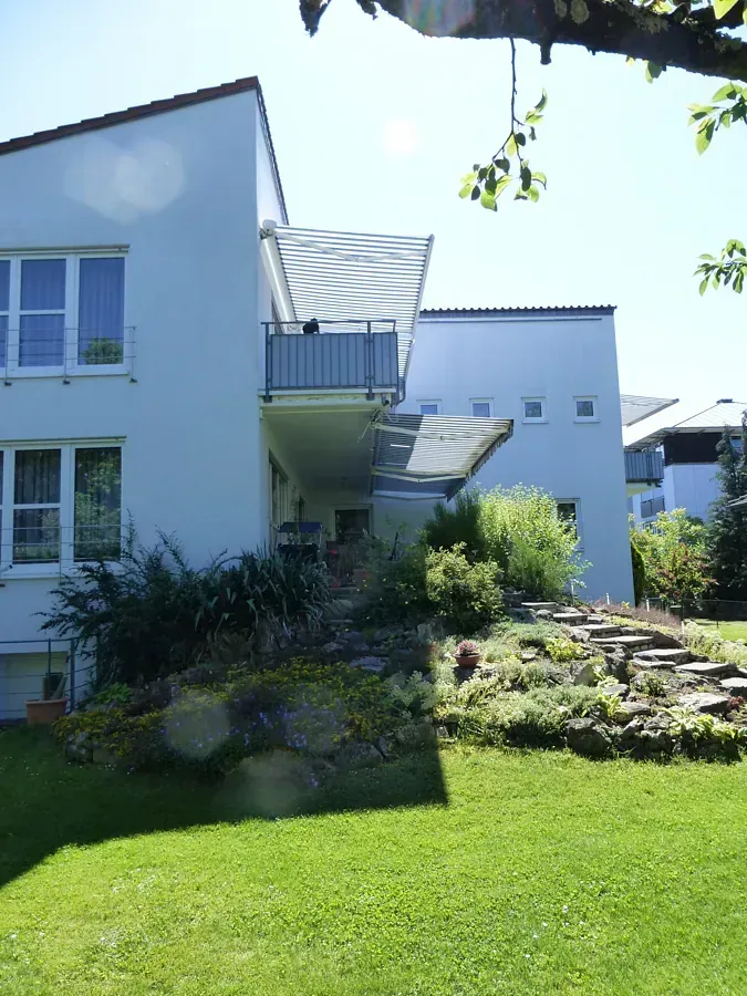 Weißes Gebäude mit Balkon und Markise über einem terrassenförmig angelegten Garten mit Treppen, von einem sonnigen Rasen aus zu sehen.