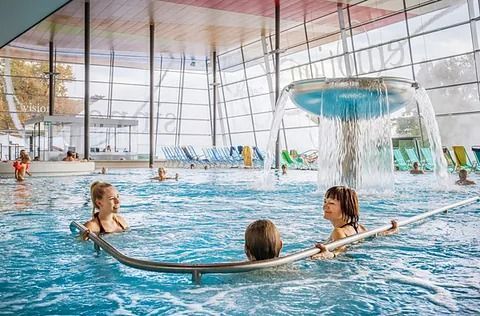Ein Hallenbad mit Badegästen; ein Springbrunnen sprudelt. Mehrere Personen befinden sich im Wasser, eine Person bedient die Bar. Blaues Wasser, helle Umgebung.