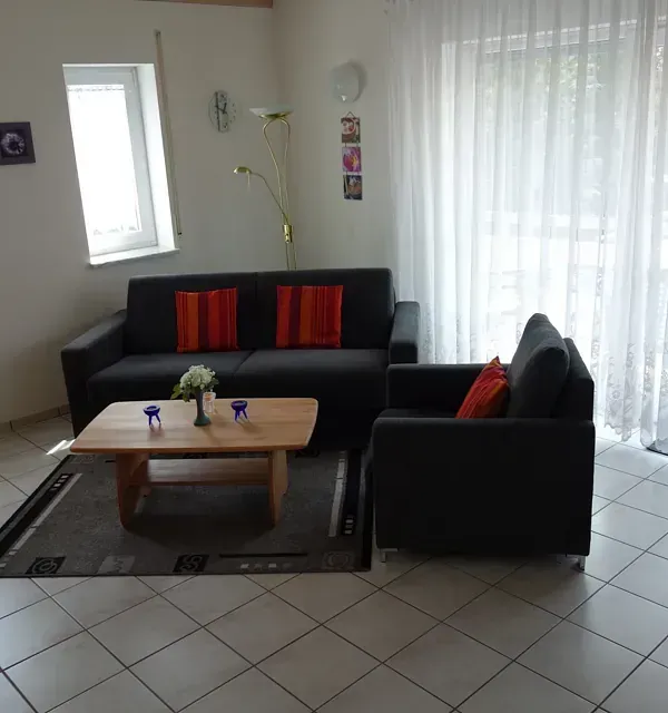 Wohnzimmer mit schwarzem Sofa, Sessel, Couchtisch und weißen, transparenten Vorhängen. Orangefarbene Kissen.