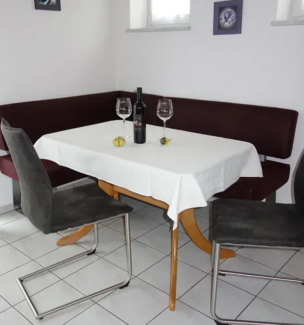 Esstisch mit Eckbank, Stühlen, Weinflasche und Gläsern. Weiße Tischdecke, Fliesenboden.
