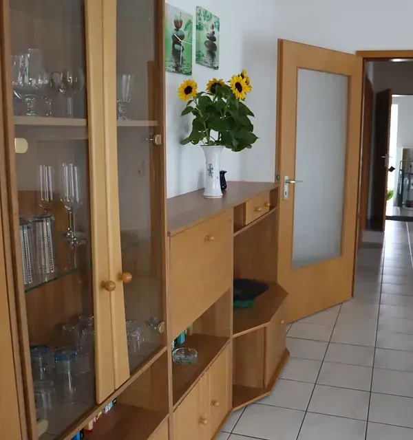 Holzschrank mit Glastüren, Sonnenblumen in einer Vase und ein Türrahmen. Helle Wände und gefliester Boden.