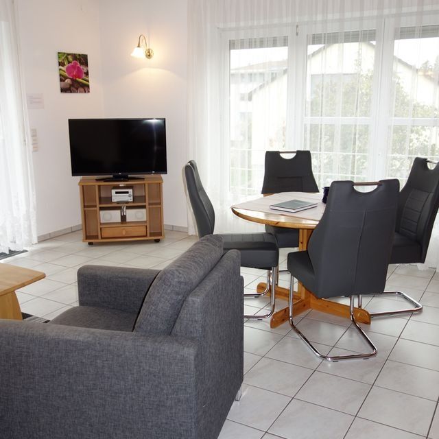 Wohnzimmer mit Fernseher, Sofa, Tisch, Stühlen und Fenster. Graue Holzmöbel, weißer Fliesenboden.