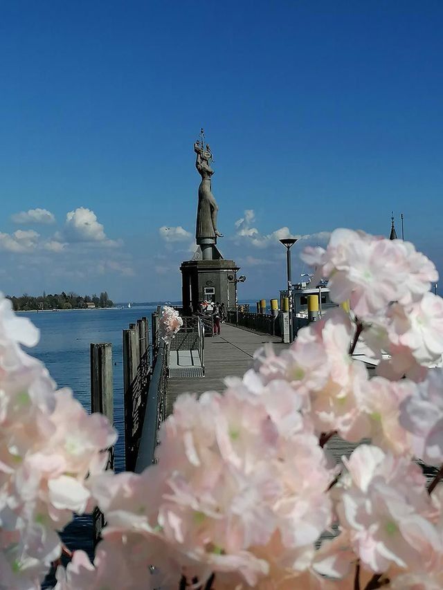 Löwenstatue und Leuchtturm auf einem Holzsteg, Blumen im Vordergrund, blauer Himmel.