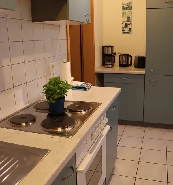 Küche mit Herdplatte, Schränken und Geräten in einem Raum mit Fliesenboden und einer Tür.