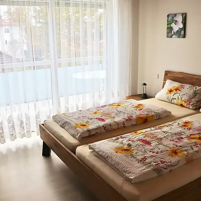 Schlafzimmer mit zwei Betten, bezogen mit geblümter Bettwäsche, neben einem Balkon mit transparenten Vorhängen.