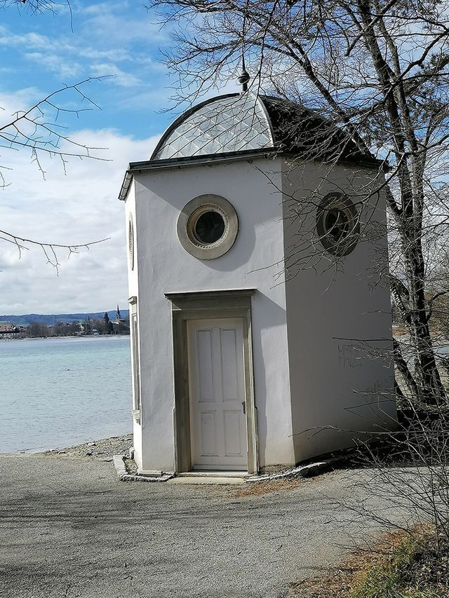 Kleines, weißes, achteckiges Gebäude mit Kuppeldach und runden Fenstern an einem See.