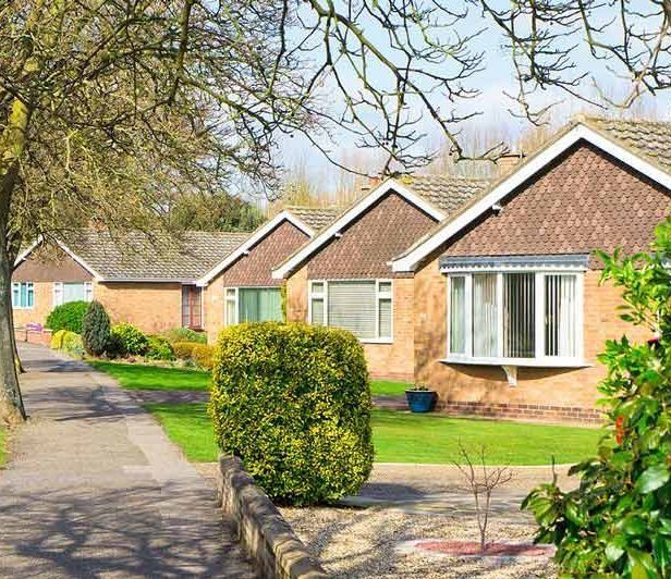 Una hilera de bungalows de ladrillo con ventanales y tejados a dos aguas, situados a lo largo de un sendero pavimentado con césped verde y arbustos.