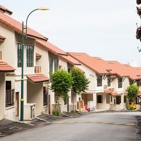 Una hilera de casas adosadas de varias plantas con paredes beige y tejados de tejas naranjas bordea una calle pavimentada en un día nublado.