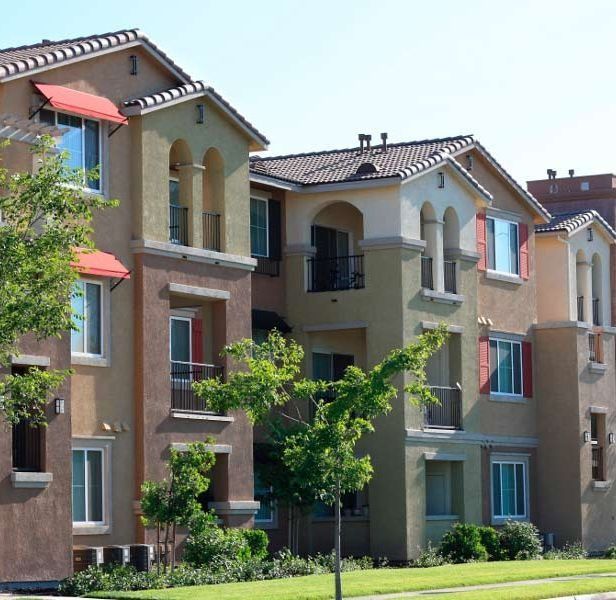 Un edificio de apartamentos de varias plantas con exterior de estuco, balcones arqueados y toldos rojos, con el cielo azul de fondo.