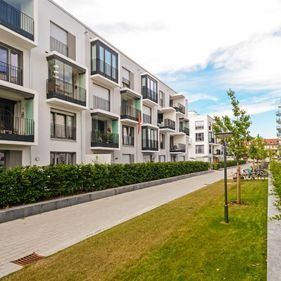 Un moderno complejo de apartamentos blancos con balcones, adyacente a un paseo pavimentado y un cuidado césped verde bajo un cielo azul.