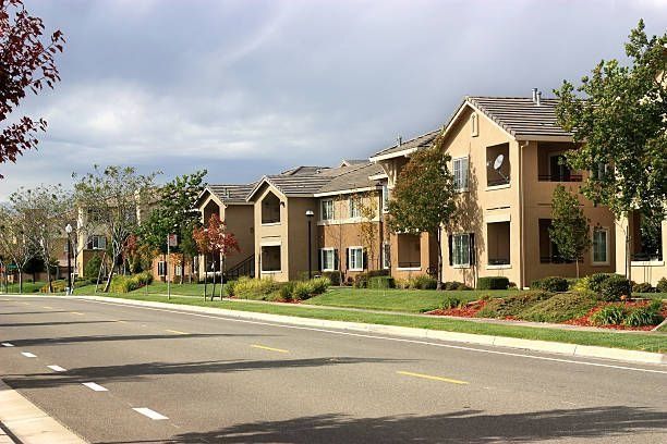 Vista de la calle con edificios de apartamentos de varias plantas de color beige y tejados a dos aguas, rodeados de césped y árboles.