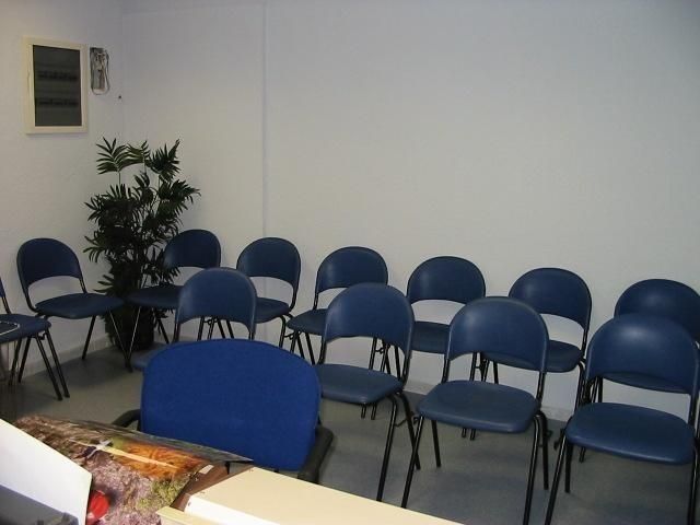 Una sala de reuniones con una fila de sillas azules frente a un escritorio, una planta en maceta en la esquina y una pared blanca.