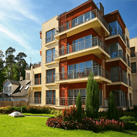 Edificio de apartamentos moderno con fachada en tonos crema y marrón, balcones y grandes ventanales con vistas a un césped.