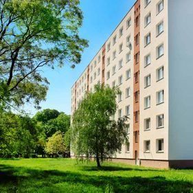 Un edificio de apartamentos alto y de color claro se alza junto a un césped arbolado bajo un cielo azul despejado.