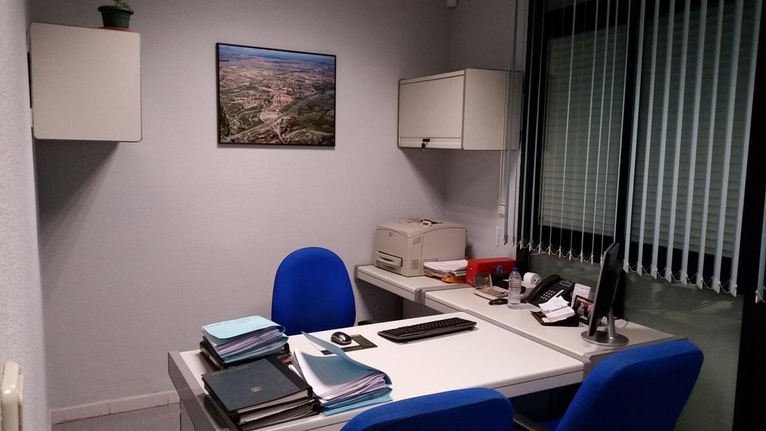 Una pequeña oficina con un escritorio con dos sillas azules, dos armarios empotrados en la pared, un ordenador y un cuadro mural con un paisaje.