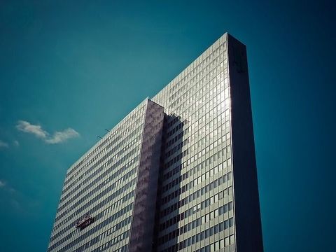 Un moderno rascacielos de cristal de líneas angulares se eleva hacia un cielo azul intenso, con una pequeña plataforma de mantenimiento visible en su fachada.