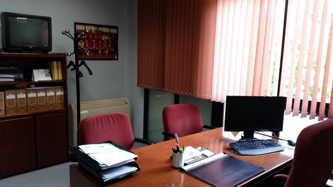 Una oficina con un escritorio de madera, tres sillas rojas, un ordenador, un televisor y estanterías con archivos bajo persianas verticales.