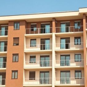 Edificio de apartamentos de varias plantas con paredes beige, columnas marrones y balcones de cristal bajo un cielo azul despejado.