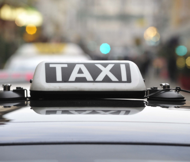 Panneau de toit de taxi avec « TAXI » en lettres blanches, monté sur une voiture noire. Ville en arrière-plan.