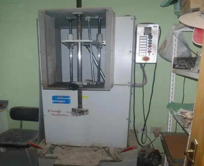 Máquina de ensayo industrial en un taller, con panel de control y accesorios metálicos dentro de un armario abierto.