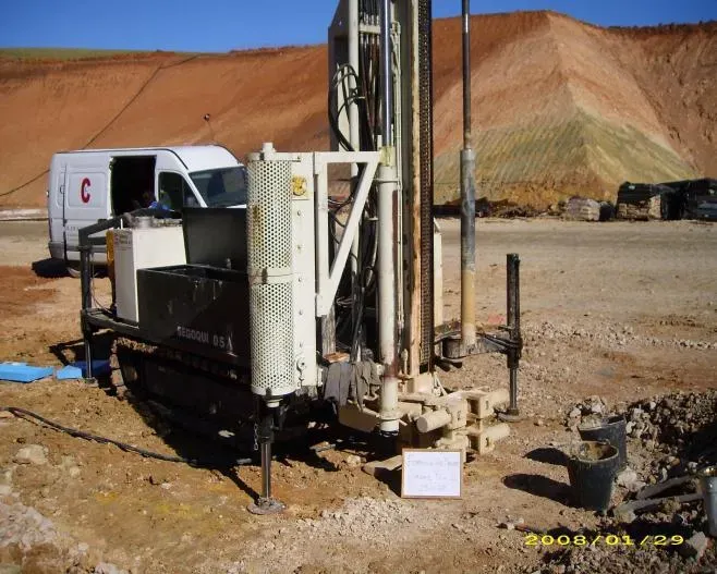 Plataforma de perforación y camión en un terreno baldío con un terraplén de roca roja al fondo.