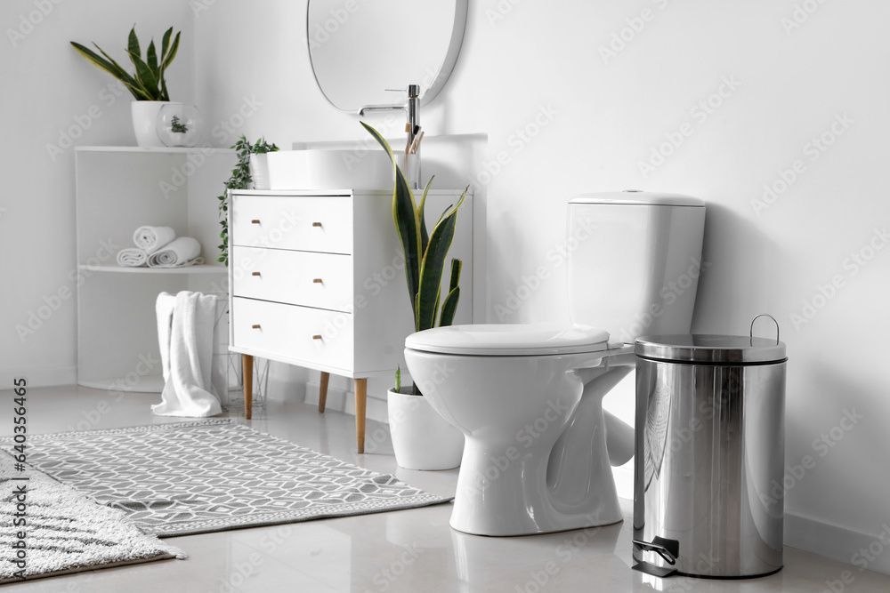 Helles, weisses Badezimmer mit Toilette, Waschbecken, Schubladen, Pflanzen und silbernem Mülleimer