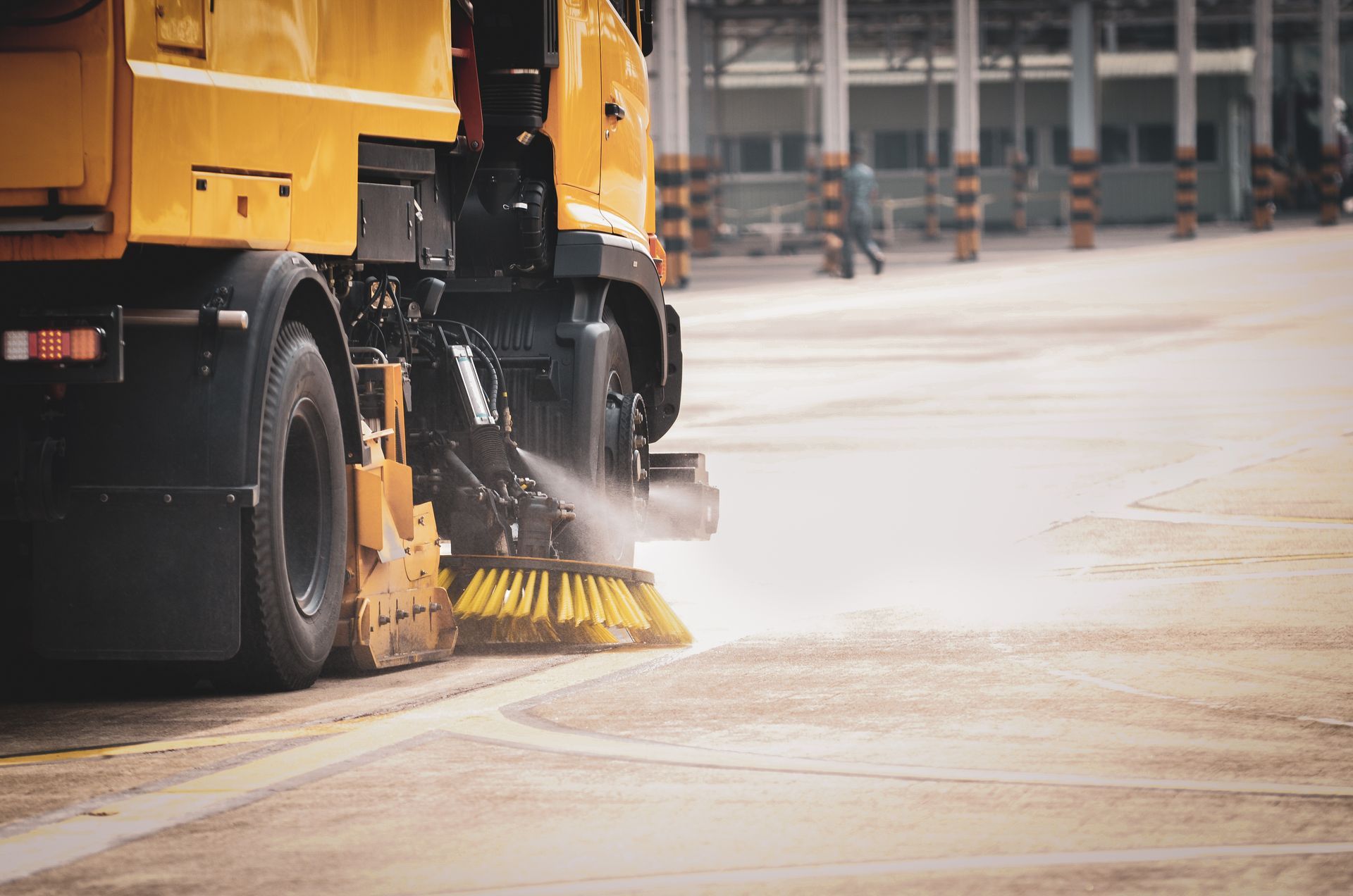 Balayeuse de rue jaune nettoyant une surface pavée ; brosses et jet d'eau en action.