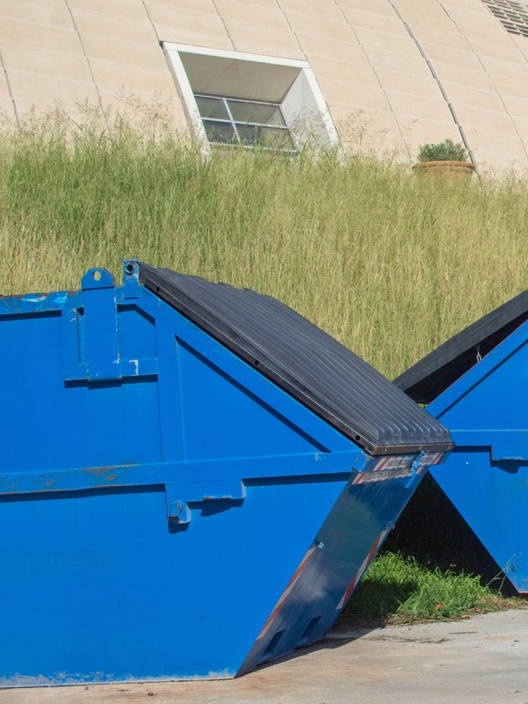 Benne bleue avec couvercle ouvert, devant de hautes herbes et un bâtiment avec une fenêtre.