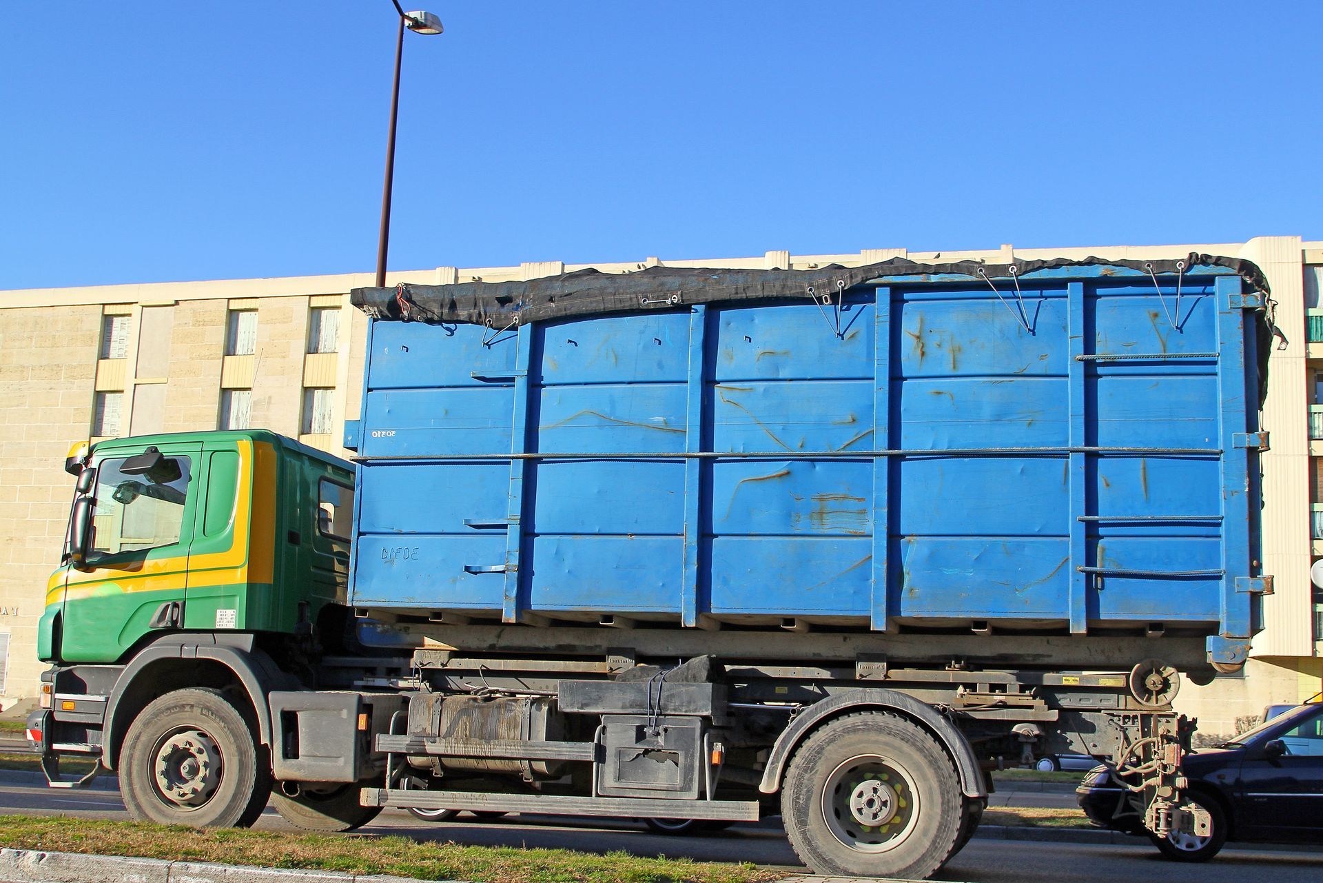 Camion d'élimination des déchets bleu avec cabine verte garé sur le bord d'une route, bâtiment en arrière-plan.
