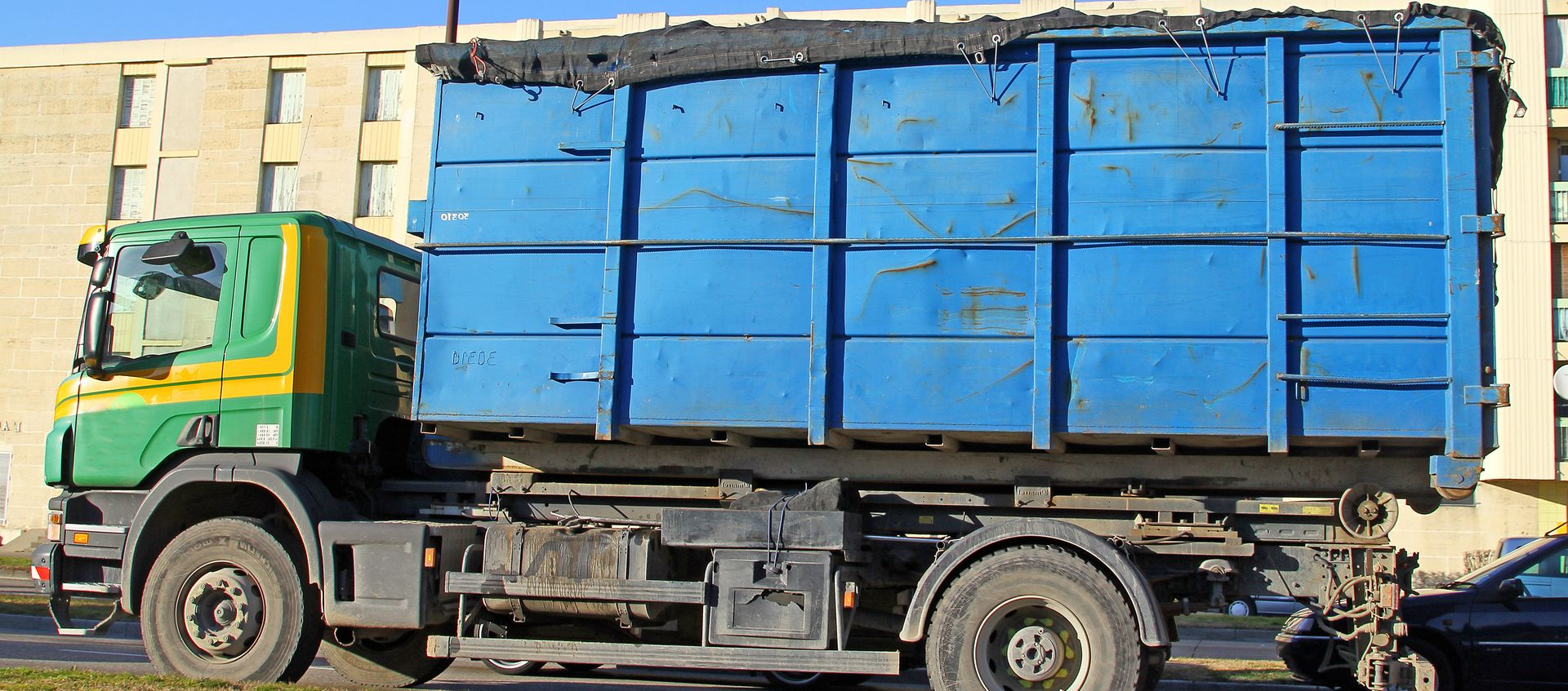Un camion-benne bleu avec une cabine verte et jaune est garé devant un bâtiment beige.