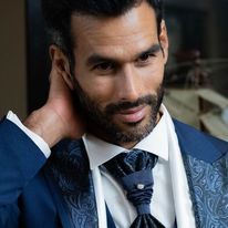 Hombre con traje azul marino, corbata de cachemira, mano en el cuello, mirando hacia abajo, entorno interior.