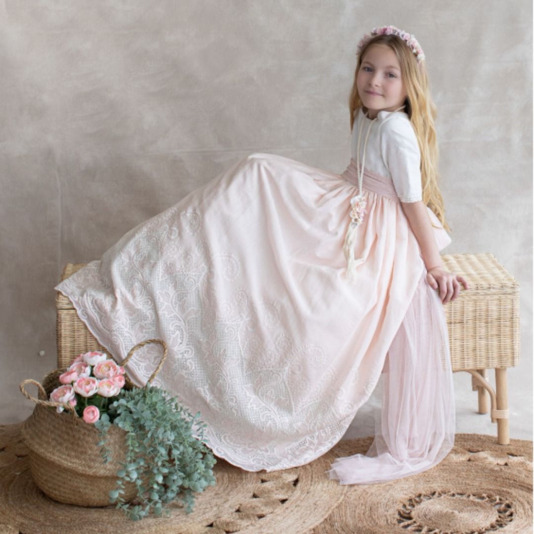 Una niña con un vestido rosa pálido y una diadema de flores está sentada en un banco de mimbre