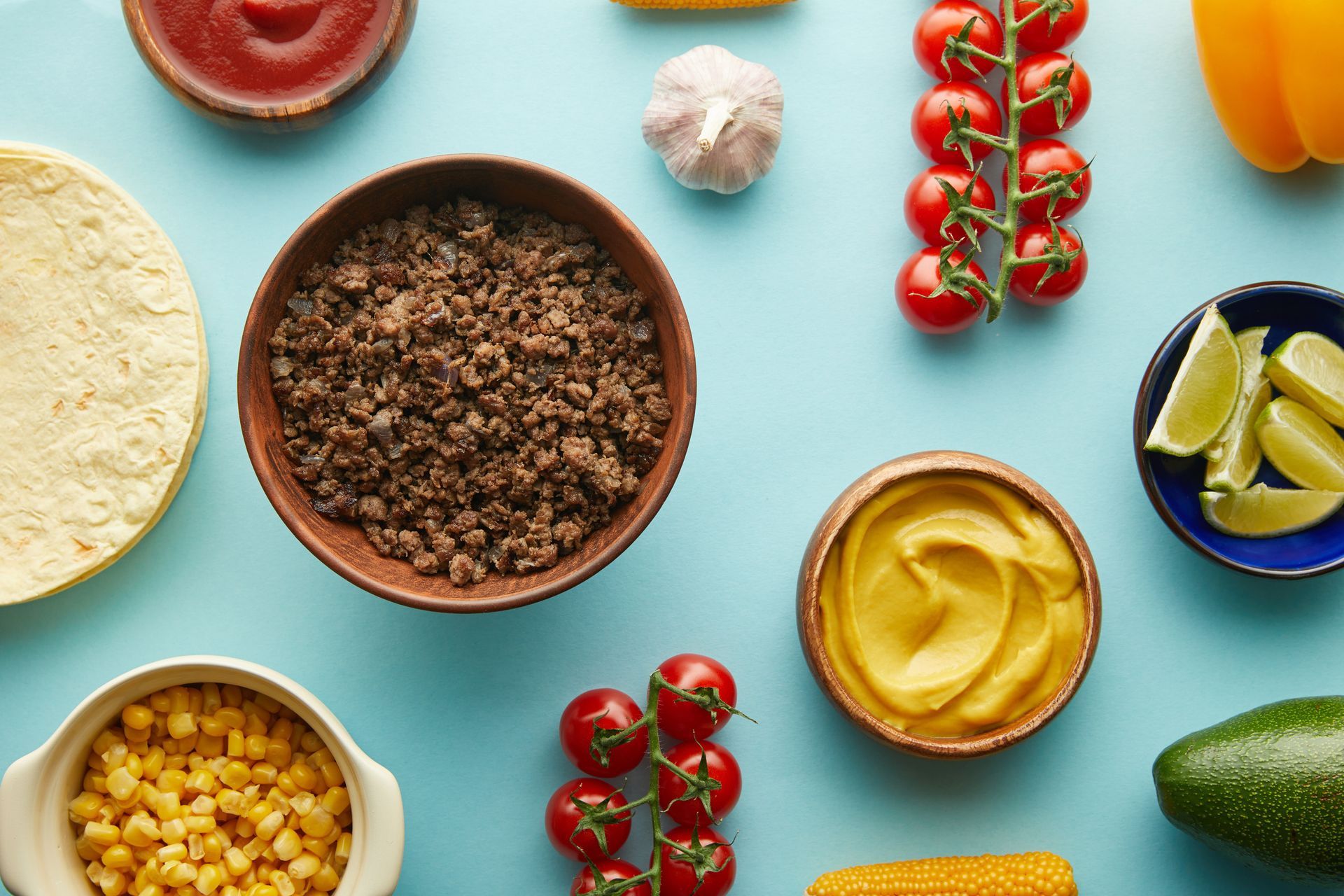 Ingredientes del taco: carne molida, tortillas, maíz, salsa, tomates, limones, aguacate y salsas, dispuestos sobre una superficie azul.