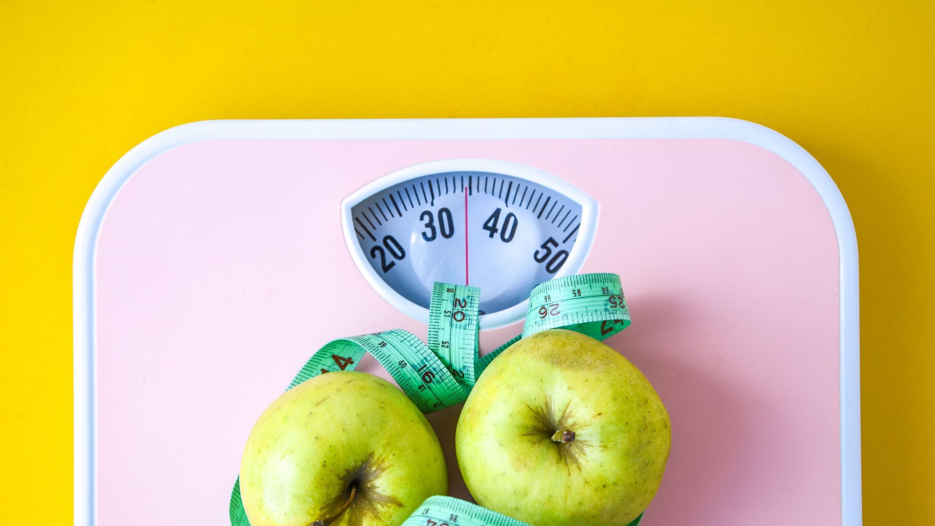 Báscula rosa con manzanas y cinta métrica, que sugiere pérdida de peso y alimentación saludable.