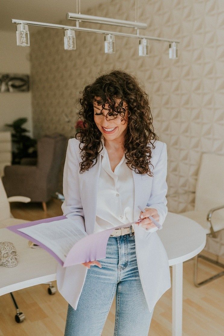 Mujer con cabello rizado en blazer y jeans, sonriendo, sosteniendo papeles cerca de una mesa blanca.