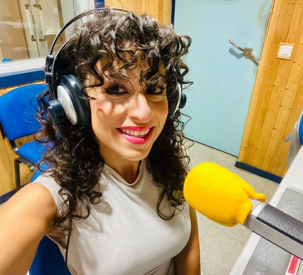 Mujer en un estudio de radio con auriculares y sonriendo a la cámara, micrófono visible.