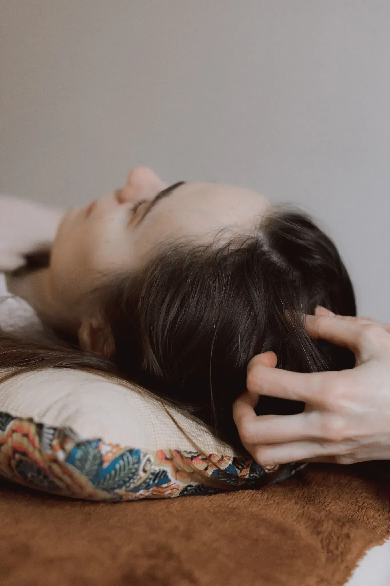 Persona recibiendo un masaje capilar, tumbada en una camilla. Masajea el cabello oscuro con la mano; expresión neutra.