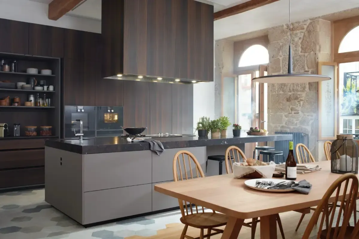 Cocina moderna con gabinetes de madera, isla y mesa de comedor; luz natural desde las ventanas.