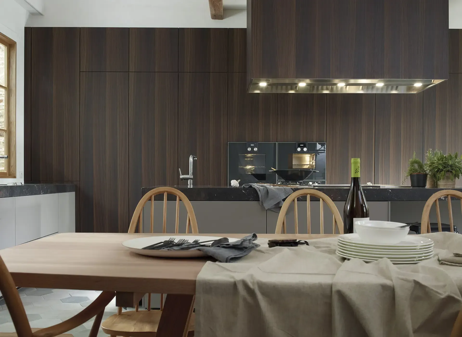 Comedor con mesa de madera preparada para comer frente a una cocina moderna con paneles de madera oscura.