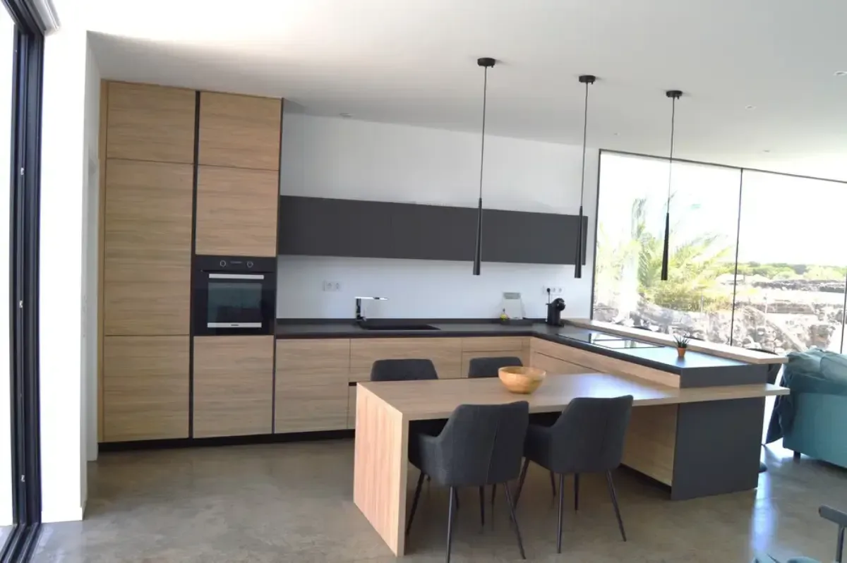 Cocina moderna con gabinetes de madera, encimeras oscuras y una mesa de comedor con sillas.