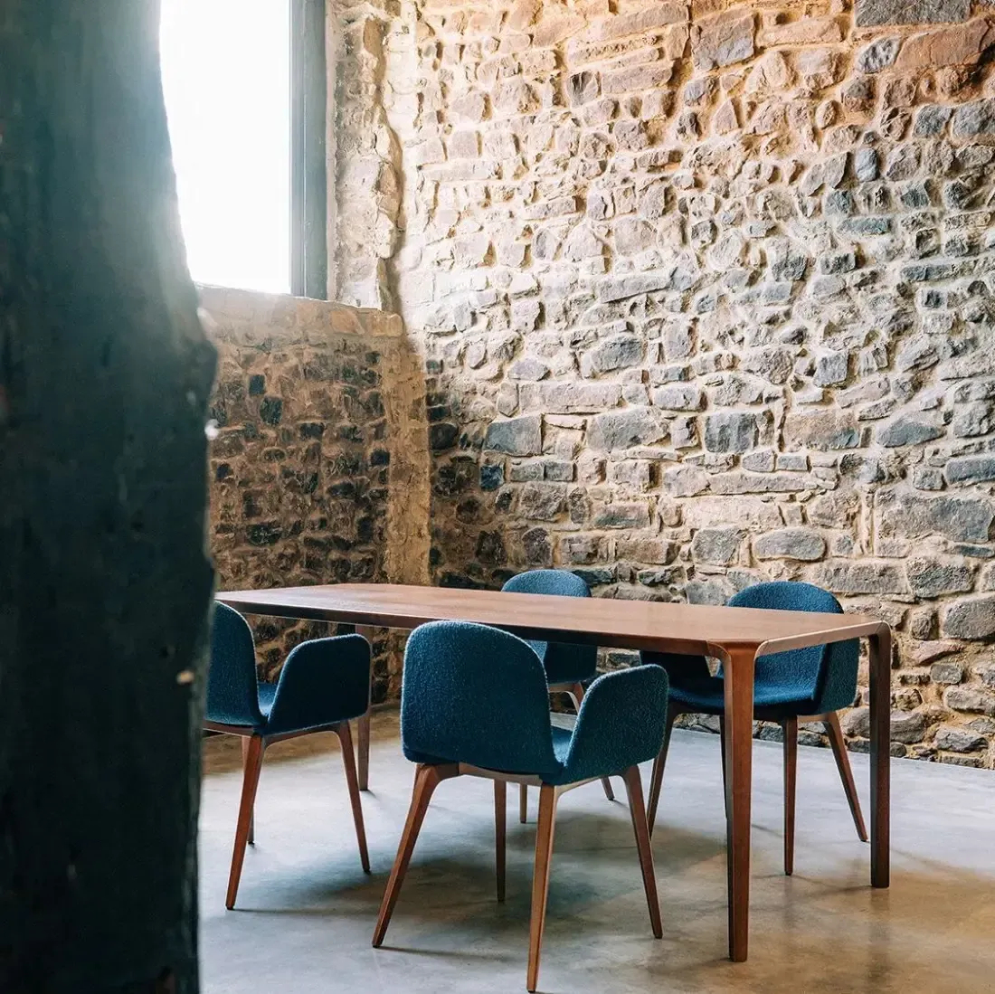 Mesa de comedor con cuatro sillas azules frente a un muro de piedra, cerca de una ventana.