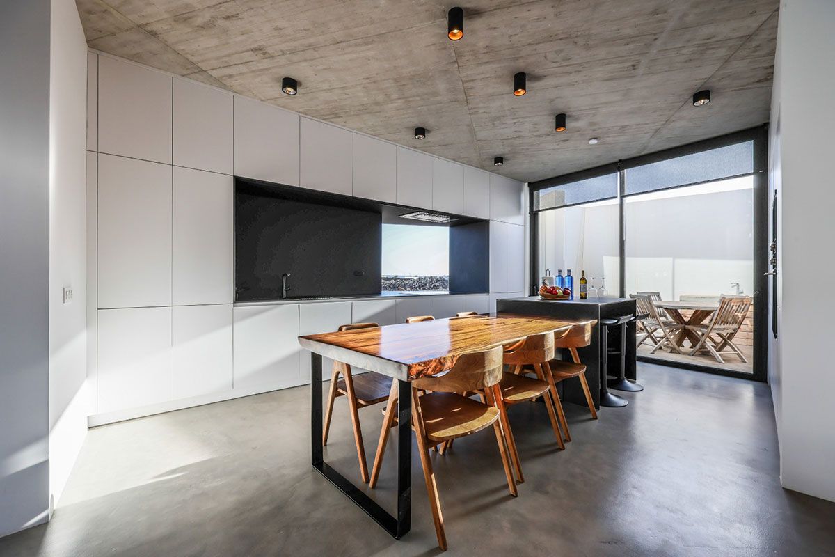 Cocina moderna con gabinetes blancos, techo de concreto, mesa y sillas de madera y una puerta de vidrio a un patio.