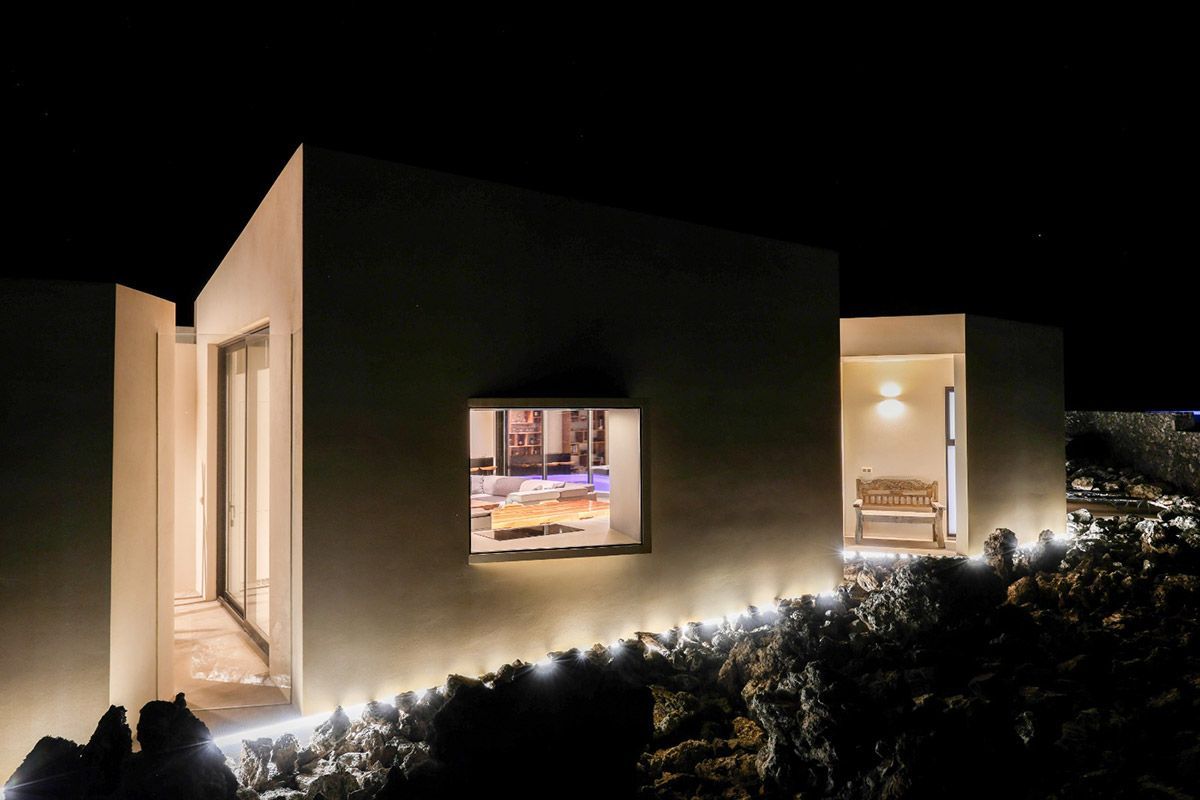 Vista nocturna de un moderno edificio blanco con luces brillantes, ventana iluminada que muestra el interior, un banco al aire libre y follaje oscuro.