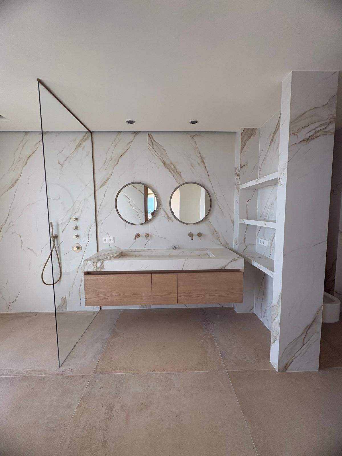 Tocador de baño moderno con espejos redondos, paredes con aspecto de mármol, armarios de madera y panel de ducha de cristal.