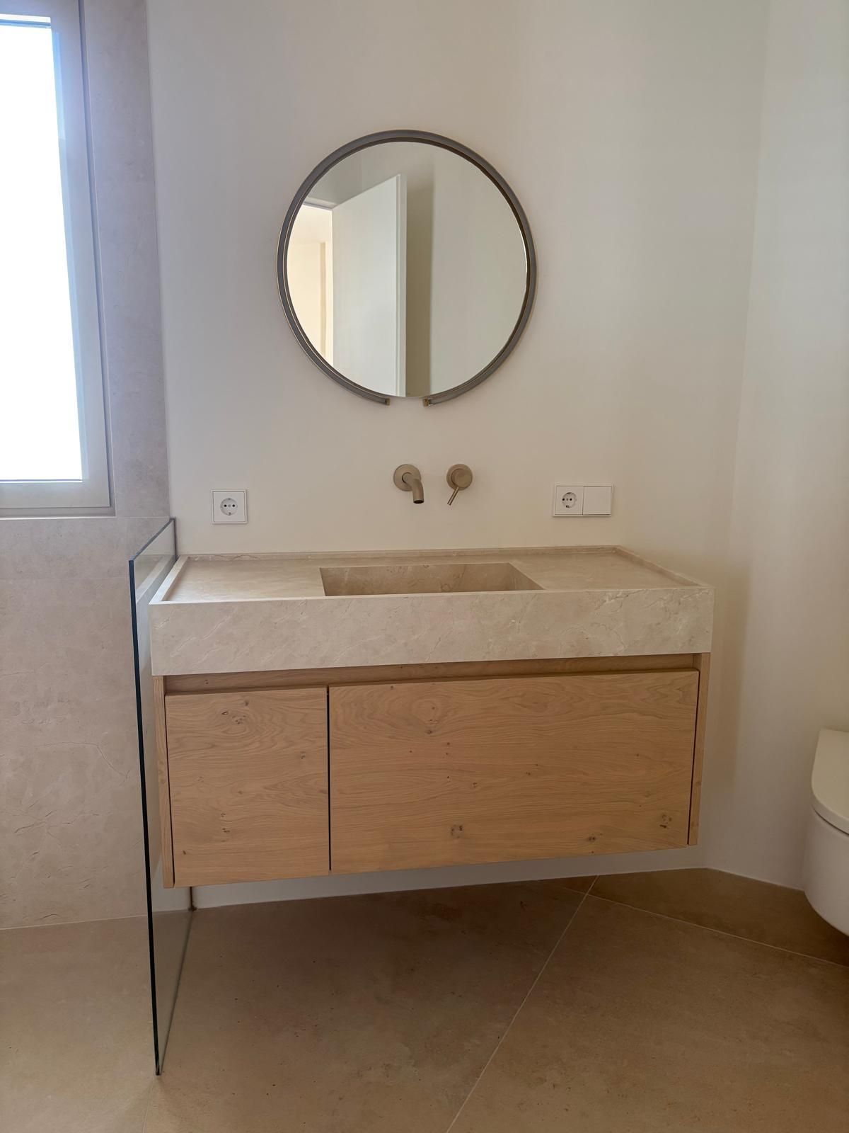 Tocador de baño moderno con espejo redondo, mueble de madera y encimera de piedra junto a una ventana.